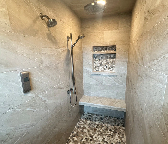 Remodeled bathroom shower with bench, three shower heads, light, and cream marble tiles on the walls and ceiling, and river rock tiles on the floor.