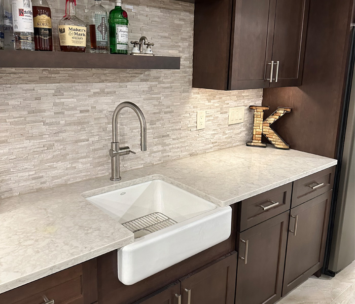 Small, under-mount, white farmhouse sink in a home bar with a light marble countertop and dark brown cabinets.