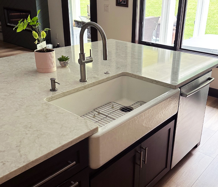 Under-mount, white farmhouse sink in a kitchen island with a light marble countertop and dark cabinets.