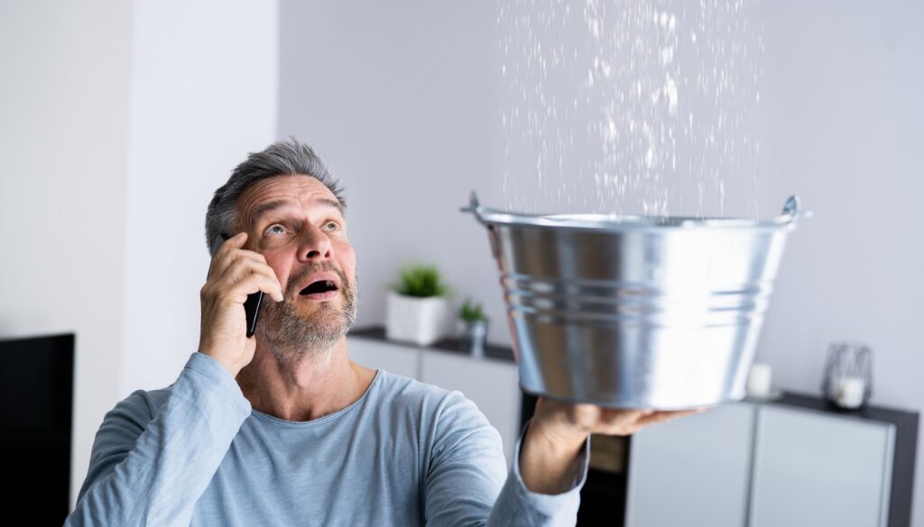 A man talking on the phone while holding a bucket to catch water dripping from the ceiling.