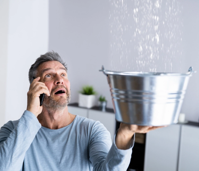 A man talking on the phone while holding a bucket to catch water dripping from the ceiling.