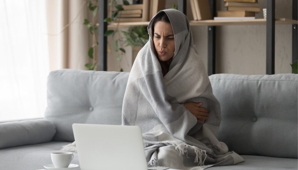 Person bundled in a blanket on a couch, looking cold and uncomfortable.