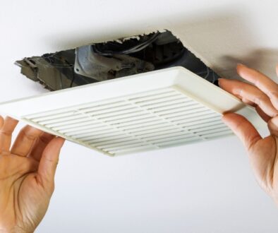 Hands installing a bathroom ventilation fan cover on a ceiling, with exposed wiring and ductwork visible in the opening.