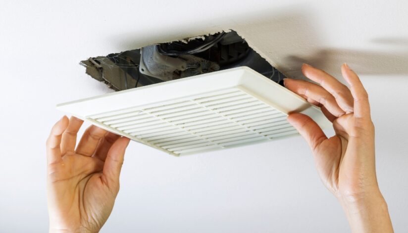 Hands installing a bathroom ventilation fan cover on a ceiling, with exposed wiring and ductwork visible in the opening.