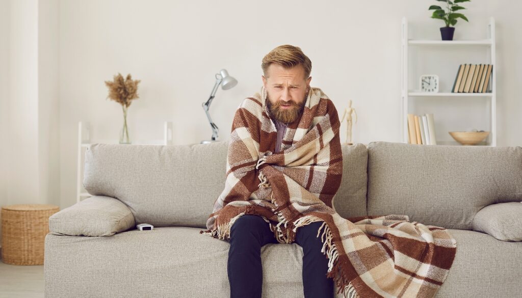 Man wrapped in a blanket sitting on a couch, looking cold and uncomfortable.