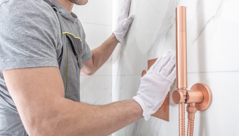A professional contractor aligning marble wall tiles during a bathroom shower remodel, featuring a modern copper shower fixture.
