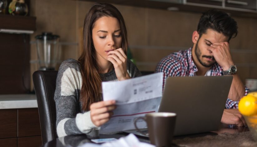 A worried couple sits at their kitchen table reviewing a high water bill, with a laptop and paperwork in front of them.