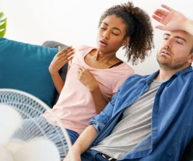 niebuhr-plumbing-heating-air-conditioning-wahy-is-one-room-hotter-than-others-couple-sitting-on-couch-in-front-of-fan