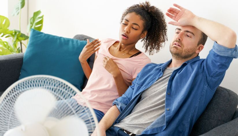 niebuhr-plumbing-heating-air-conditioning-wahy-is-one-room-hotter-than-others-couple-sitting-on-couch-in-front-of-fan