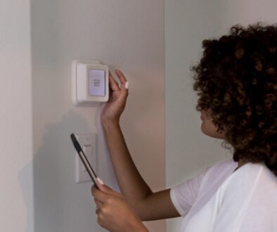 A woman adjusting a wall-mounted thermostat set to 69 degrees while holding a smartphone to test her furnace before winter.