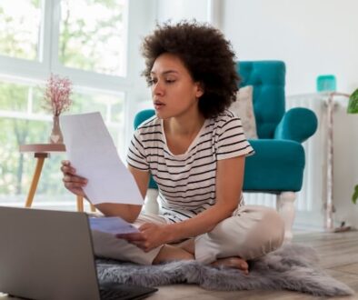 A woman reviewing a utility bill while sitting on the floor with a laptop.