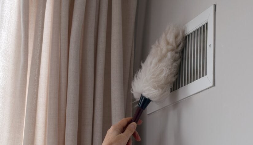 A hand using a feather duster to clean dust from a wall air vent next to beige curtains.